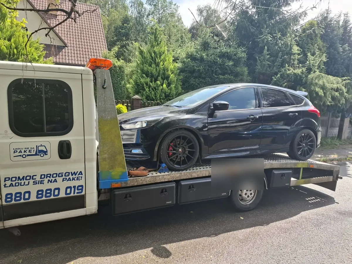 Czarny Ford Focus na platformie lawety przy domach